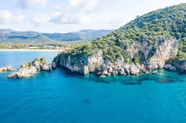 Parlak gün ışığı, Yunanistan 'ın Epirus kentindeki Karavostasi plajının turkuaz suları boyunca yemyeşil bitki örtüsüyle kaplı engebeli kayalıkları ve kayalık adaları aydınlatıyor. Sahne, açık mavi deniz ve uzak Akdeniz kıyılarını yakalıyor.