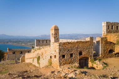Yıpranmış kale duvarları ve mavi suların üzerinde yuvarlak bir gözetleme kulesi ve Yunanistan 'ın Nafplio kentindeki Palamidi Kalesi' nde uzak dağlar. Ilık güneş ışığı, dokulu duvarcılık ve engebeli manzarayı vurgular.