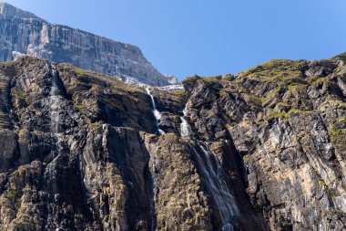 Dağ manzarasının dramatik dokularını ve doğal katmanlarını vurgulayan parlak mavi gökyüzünün altında, Gavarnie Sirki 'ndeki sarp ve kayalık bir uçurumdan birkaç su akıntısı akar..