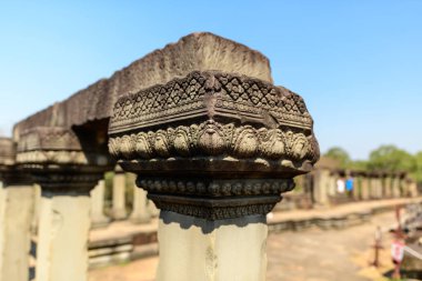 Angkor, Kamboçya 'daki Baphuon Tapınağı' nın zengin kumtaşı sütun başkentinin yakın çekim görüntüsü. Güneş ışığı, arka planda bulanık harabeler ve mavi gökyüzü ile ayrıntılı Khmer motifleri ve desenli taş vurgular.