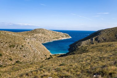 Yunanistan 'ın Cap Tenare kentindeki engebeli, güneşli tepeler arasında gizli bir turkuaz koy bulunuyor. Canlı mavi su, berrak bir gökyüzünün altındaki kuru Akdeniz bitki örtüsüyle çelişiyor ve el değmemiş bir sahil güzelliği hissi uyandırıyor..