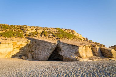 Katmanlı kumtaşı mağarası ve kayalık çıkıntısı Triopetra plajı, Girit 'te parlak mavi gökyüzü altında dokulu uçurumlar ve seyrek yeşilliklerle güneşlenir. Sahne sıcaklığı, sertliği ve doğal güzelliği çağrıştırıyor..