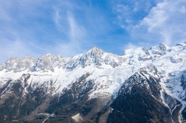 Aiguille du Midi 'nin sarp zirvesi, karla kaplı yamaçlar ve dinamik mavi gökyüzünün altındaki dağınık ormanlarla birlikte Bossons Buzulu' nun üzerinde yükseliyor..
