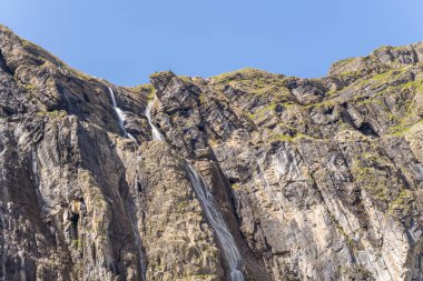 Çok ince şelaleler, berrak mavi bir gökyüzü altında, keskin dokuları ve yeşil parçalarıyla, Gavarnie Sirki 'nde engebeli, güneşli kaya yüzeylerine inerler. Sahne dramatik yükseliş ve doğal dağ jeolojisini vurguluyor.