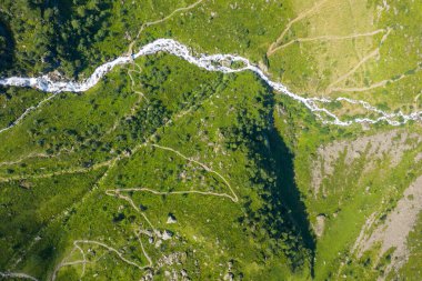Dar bir beyaz su nehrinin yüksek açılı drone görüntüsü yemyeşil yamaçlar boyunca kıvrılıyor ve Pyrenean kırsalındaki kayalık arazide, patikalar ve dağınık kayalar ile benekli..