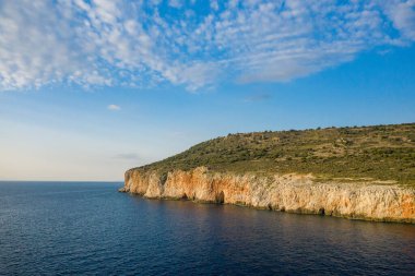Altın uçurumlar, yumuşak bulutlarla kaplı bir gökyüzünün altında uzanan yumuşak yeşil yamaçlarla, sakin derin mavi suların üzerinde yükselir. Sahne, sıcak güneş ışığıyla aydınlanıyor, doğal dokuları ve kıyı huzurunu vurguluyor..