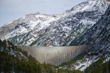 İtalya 'nın Aosta Vadisi' ndeki Place-Moulin yakınlarındaki engebeli, karlı dağlara ve yemyeşil ormanlara kurulmuş devasa beton barajın geniş bir manzarası. Kış manzarasında desenli kaya yüzeyleri, dağınık çam ağaçları ve çıtır çıtır yüksek irtifa yer alır.