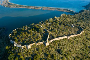 Yunanistan 'ın Navarino kenti yakınlarındaki mavi kıyı lagününün üzerindeki engebeli, yeşil bir tepenin üzerinde korunmuş duvarları olan geniş bir ortaçağ taş kalesinin havadan çekilmiş fotoğrafı, altın güneş ışığıyla yıkanmış..