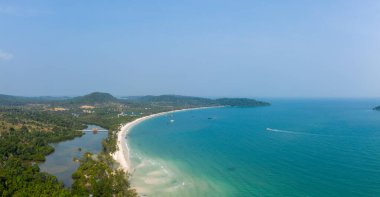 Panoramik hava manzarası, Kamboçya 'nın Koh Rong adasındaki Long Set Sahili' nin geniş kıvrımlarını gösteriyor. Yoğun orman, yumuşak tepeler ve dağınık tekneler berrak mavi gökyüzünün altındaki bu sakin tropikal kıyı şeridini tanımlar..