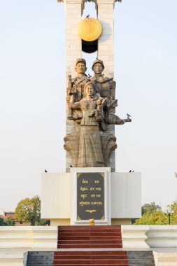 Kamboçya 'nın Phnom Penh kentindeki bir platformun üzerinde üç stilize figürün yer aldığı heykel duruyor. Ilık sabah ışığı, yapı üzerinde güvercinler tünemiş ve arka planda açık gökyüzü ile desenli taş ve altın madalyonu vurgular.