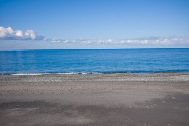 Plage de Acchio, Fiumicello 'da kumlu ve çakıl taşlı kıyı şeridi yumuşak bulutlarla benekli parlak bir gökyüzü altında sakin mavi denizle buluşan dalgalar..