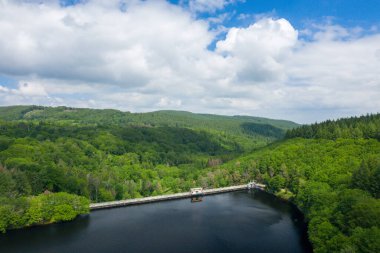 Yoğun, hareketli ormanlarla ve Chaumecon gölünde dramatik bulutlarla dolu bir gökyüzünün altındaki dalgalı tepelerle çevrili derin ve sakin bir göl boyunca beton bir baraj uzanır..