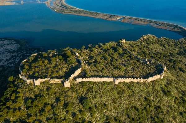 Yunanistan 'ın Navarino kenti yakınlarındaki sakin mavi bir lagünün üstündeki yemyeşil, ormanlık bir tepenin üzerinde süzülen antik taş kale duvarlarının havadan perspektifi, gün ışığı altında.