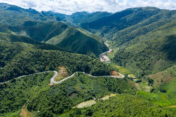 Laos 'taki Phou Khoun ve Luang Prabang arasında yoğun yeşil tepeler ve vadilerden geçen eğimli bir dağ yolunun hava manzarası. Manzarada katmanlı tepeler, dağınık kırsal binalar ve dramatik bulut gölgeleri yer alıyor.