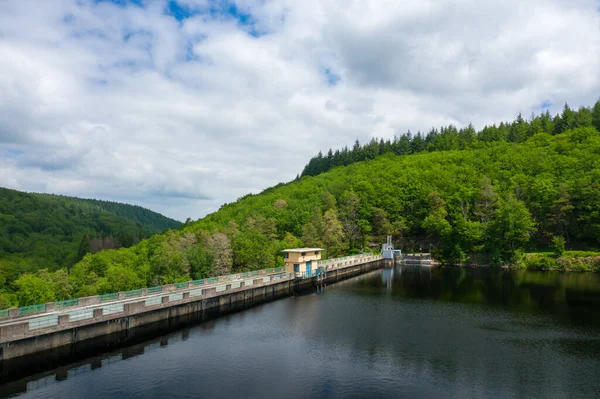 Sari bir kamusal bina, gölgelik sulara bakan beton bir barajın üzerinde duruyor. Etrafi yemyeşil orman tepeleriyle çevrili, Chaumecon gölünde bulutlu bir gökyüzünün altında..