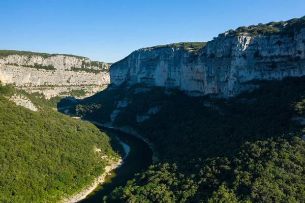 Kapsamlı bir hava manzarası, Gorges de lArdeche 'deki kıvrımlı bir nehrin altında gür ve yoğun bir orman vadisinin üzerinde yükselen dramatik kireçtaşı uçurumlarını ortaya çıkarıyor. Engebeli arazi parlak mavi gökyüzünün altında berrak güneş ışığıyla yıkanır..