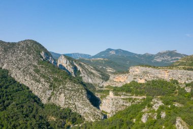 Geniş kireçtaşı çıkıntıları ve yeşil orman yamaçları, açık mavi gökyüzünün altındaki uzak dağlarla Verdon Boğazı 'nın dramatik manzarasını tanımlar..