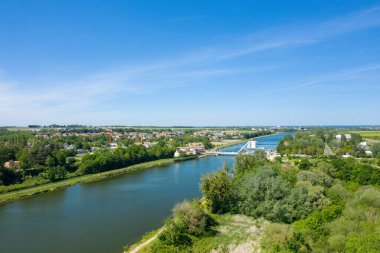 Bereketli yeşillikler, köy çatıları ve Normandiya 'daki canlı mavi gökyüzünün altındaki açık kırsal alanda Pegasus Köprüsü' nün sakin bir nehri geçişini gösteren geniş bir insansız hava aracı görüntüsü..