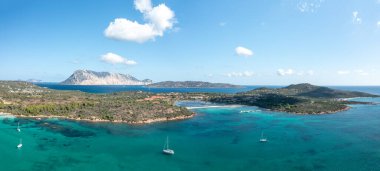 San Teodoro, Sardinya yakınlarındaki Spiaggia Salina Bamba 'nın hava manzarasında turkuaz sular, demirlemiş yelkenliler, engebeli kıyı şeridi ve Tavolara adasının çarpıcı silueti parlak mavi gökyüzü altında yer almaktadır..