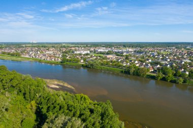 Sully-sur-Loire 'nin açık mavi gökyüzünün altında yeşil ağaçlar ve yerleşim çatıları ile Loire Nehri' nin kasabayı kıvrımlı şekilde geçişini gösteren geniş bir hava manzarası. Manzara açık, canlı ve huzurlu..