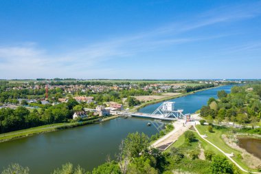 Pegasus Köprüsü 'nü canlı yeşillikler, köy çatıları ve Normandiya' da açık mavi bir gökyüzünün altındaki açık arazilerle çevrili sakin bir nehri geçerken gösteren hava manzarası..