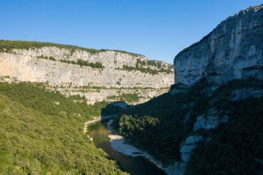 Kavisli bir nehrin yukarısındaki dramatik kireçtaşı uçurumları Gorges de Ardeche 'deki yoğun yeşil ormanlarla çevrili. Parlak güneş ışığı ve derin gölgeler manzaranın engebeli dokularını ve doğal hatlarını güçlendirir..