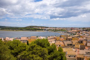 Gruissan köyünün güneşli terracotta çatılarında geniş bir lagün ve bulutlu bir yaz gökyüzünün altında uzak kıyı şeridi olan yüksek perspektif. Yeşil yemyeşil ağaçlar ön planı çerçeveleyerek, Akdeniz manzarasına doğal doku ekliyor.