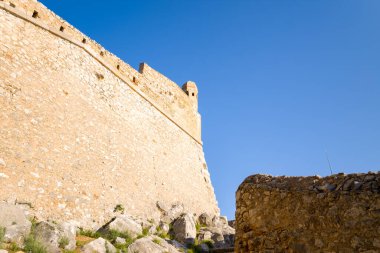 Parlak güneş ışığı Yunanistan 'ın Nafplio kentindeki Palamidi Kalesi' ndeki dokulu taş duvarları ve engebeli kayaları vurguluyor. Sahnede sıcak toprak tonları, keskin gölgeler ve parlak mavi bir gökyüzü yer alıyor..