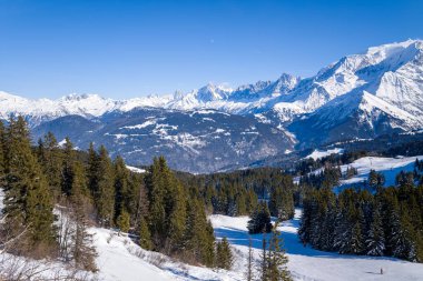 Yoğun yeşil orman battaniyeleri, parlak mavi gökyüzünün altında Mont Blanc 'ın geniş manzaralı karlı tepeleri yuvarlıyor. Parlak kış güneşi dramatik alp manzarasını ve canlı atmosferi güçlendirir..