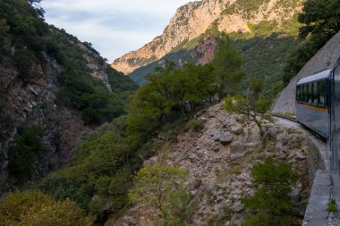 Yunanistan 'ın Kalavrita kenti yakınlarındaki Vouraikos Vadisi' nde kayalık yamaçlar ve yoğun yeşil ağaçlar boyunca dar çaplı bir tren esiyor. Öğleden sonra ışığı, bu manzaralı dağ demiryolunun dramatik kireçtaşı uçurumlarını ve engebeli arazisini vurguluyor..