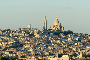 Sacre-Cur Bazilikası tarafından taçlandırılan Montmartre 'nin geniş manzarası, güneşli Paris çatıları ve berrak bir gökyüzü altında yumuşak yeşillikler, sakin bir şehir atmosferini çağrıştırıyor..
