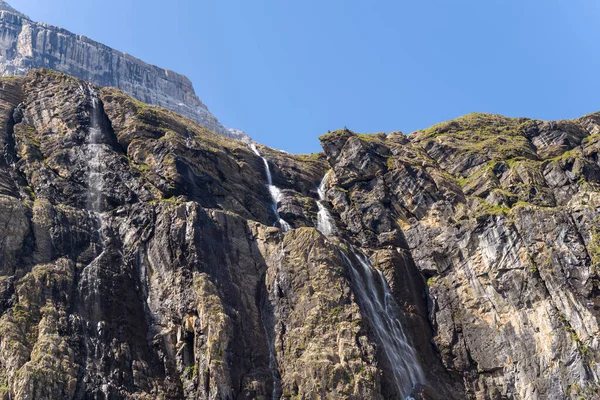 Birkaç ince şelale, açık mavi gökyüzünün altında dokulu kaya yüzeyleri ve yeşil parçalarla Cirque de Gavarnie 'deki engebeli, güneşli bir uçurumdan aşağı akıyor. Dramatik dağ manzarası doğal erozyon ve yüksekliği gösteriyor..