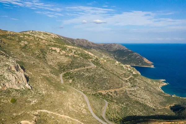 Yunanistan 'ın Magne kentindeki canlı mavi Akdeniz manzaralı güneşli kayalık tepelerin arasından bir yılan yolu geçiyor. Engebeli arazide seyrek bitki örtüsü, dokulu yamaçlar ve berrak bir gökyüzü altında geniş kıyı manzaraları bulunur..