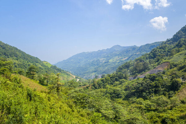 Expansive view of a lush green valley surrounded by forested hills and distant mountains in northern Vietnam. Bright sunlight highlights the vibrant foliage and creates a serene, open landscape.