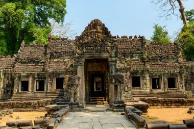 Preah Khan Tapınağı 'nın yıpranmış taş cephesi Kamboçya Angkor bölgesinde berrak bir gökyüzünün altında duruyor. Karmaşık oymalar, başsız muhafız heykelleri ve açık pencereler yemyeşil ağaçlar ve kuru otlarla çevrili doğal gün ışığıyla vurgulanır..