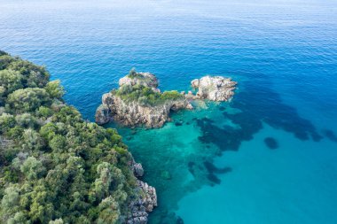 Yunanistan 'ın Epirus kentindeki Karavostasi kıyı şeridi boyunca engebeli kayalık adaları ve yemyeşil bitki örtüsünü berrak turkuaz su kaplar. Parlak gün ışığı, canlı Akdeniz sahili ve doğal kıyı şeridini vurguluyor.