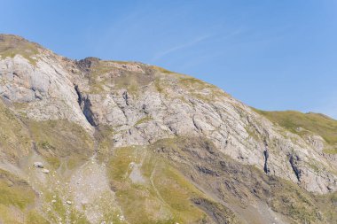 Fransız Pirenesi 'nde Gavarnie Gedre yakınlarında berrak mavi bir gökyüzünün altında kayaların tepeleri ve dağınık çimenlik alanları olan engebeli bir dağ yamacı aydınlatılır. Sahne zıt dokuları ve doğal toprak tonlarını vurgular.