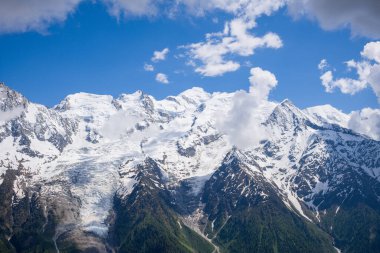 Karla kaplı zirveler ve genişleyen buzullar Mont Blanc kitlesine hükmediyor. Dağılmış bulutlar engebeli dağların üzerindeki canlı mavi gökyüzüne derinlik katıyor..
