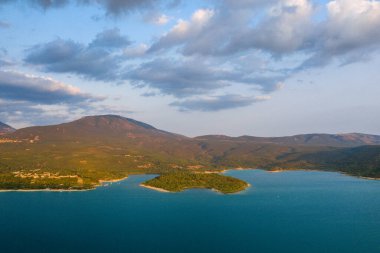 Sainte-Croix Gölü 'nün canlı turkuaz sularına uzanan sakin bir yarımada, yuvarlanan orman tepeleri ve dağınık bulutların altında akşam güneşiyle çevrelenir..