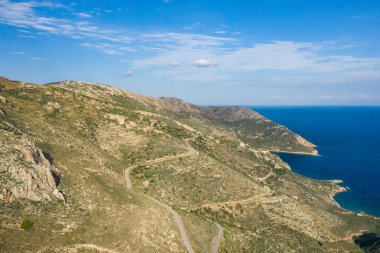 Yunanistan 'ın Magne kentindeki derin mavi denizin üzerinde kayalık tepeleri aşan dolambaçlı bir yol perspektifi. Güneşli manzarada engebeli dokular, seyrek bitki örtüsü ve parlak bir gökyüzü altında geniş kıyı manzaraları yer alır..