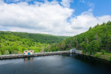 Sakin bir gölü çevreleyen beton bir barajın üzerinde küçük bir kamu binası duruyor. Etrafı yoğun yeşil ormanlarla çevrili ve Chaumecon gölündeki bulutlu mavi gökyüzünün altında yuvarlanan tepeler..