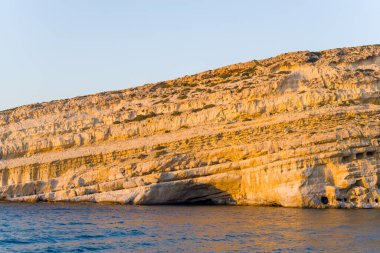 Girit 'in Matala kentindeki mavi denizin üzerinde öğleden sonra güneşiyle aydınlanan katmanlı kireçtaşı kayalıkları yükseliyor. Kayalık oluşumlar desenli yüzeyler ve doğal mağaralar sergiliyor, dramatik bir kıyı manzarası yaratıyor..
