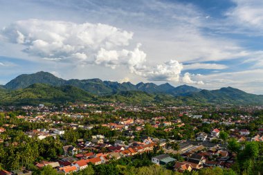 Kuzey Laos 'taki Luang Prabang' a geniş bir manzara, yemyeşil tepeler ve dramatik kümülüs bulutlarıyla çevrili kırmızı çatılardan oluşan bir demet sunar. Canlı tropikal şehir manzarası, parlak bir gökyüzünün altındaki ormanlı dağ zeminine kurulmuştur..