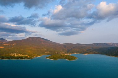 Sıcak akşam güneşi, arka planda dalgalı dağlar ve dağınık bulutlarla Sainte-Croix Gölü 'nün canlı mavi sularına giren yeşil bir yarımadayı vurguluyor..