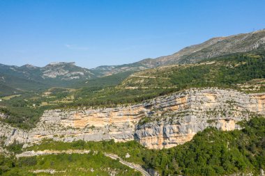 Geniş kireçtaşı kayalıkları belirgin yatay katmanlarıyla Gorges du Verdon 'daki kıvrımlı bir dağ yolunun üzerinde yükseliyor, yemyeşil tepeler ve berrak mavi bir gökyüzünün altındaki uzak tepelerle çevrili..