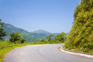 Dolambaçlı bir asfalt yol, Kuzey Vietnam 'da açık mavi bir gökyüzünün altında yoğun yeşil tepeler ve orman yamaçları boyunca kıvrılır. Güneşli yapraklar ve uzak tepeler canlı ve davetkar bir manzara yaratır..