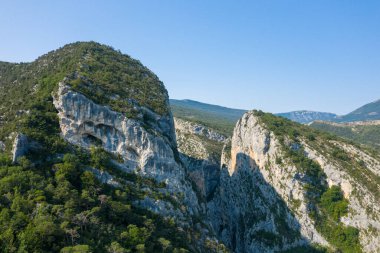 Verdon Boğazı 'ndaki yoğun yeşil ormanın üzerinde doğal mağaraları olan yüksek kireçtaşı kayalıklar, açık mavi gökyüzü ve uzak dağ sırtları altında yükseliyor..