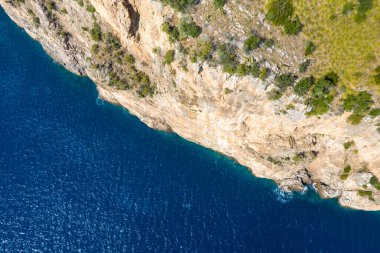 Dikey dik kayalık uçurumların derin mavi sularla buluşması, seyrek bitki örtüsü ve güneş ışığı dokularının dramatik kıyı şeridine vurgu yapması. Görüntü, doğal kaya yüzeyi ile parıldayan deniz arasındaki canlı kontrastı yakalar..