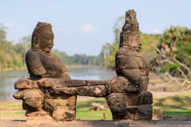 Kamboçya 'da, Siem Reap yakınlarındaki Angkor Thom' un girişinde iki antik taş koruyucu heykel balkonun üzerinde duruyor. Yıpranmış heykeller, yumuşak gün ışığında sakin bir hendek ve yemyeşil çimenlikle çerçevelenmiş ve tarihi Khmer 'i vurguluyor.