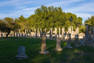 Yunanistan 'ın Olympia kentindeki olgun ağaçların altında antik Filistin' den kalma yontulmuş taş sütunlar duruyor. Öğleden sonra saatlerinde sıcak güneş ışığı arkeolojik alanda uzun gölgeler oluşturarak harabelerin ve harabelerin sakin karışımının altını çiziyor.